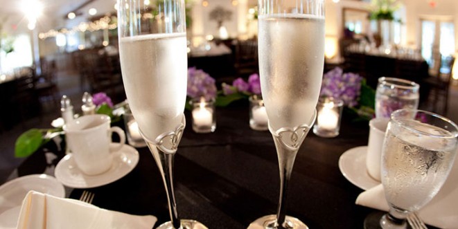 Two champagne flutes on a decorated black table with glasses, candles, and purple flowers.