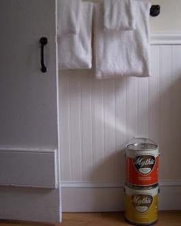 Two cans of red and yellow paint sitting on the floor of a room with white paneling, white door, and white towels.