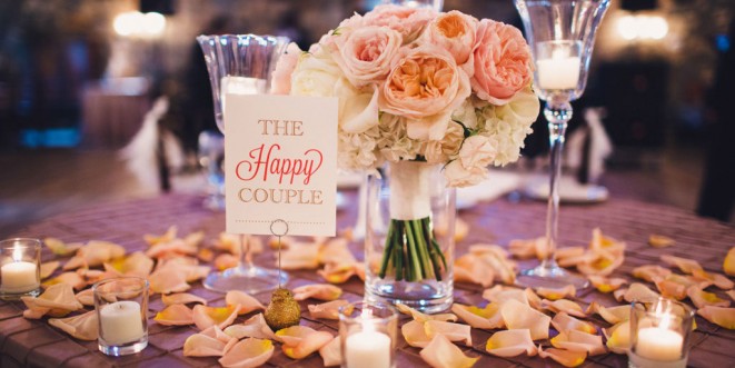 A small vase with a pink and white rose bouquet on a candle-lit table; "The Happy Couple" sign adjacent.