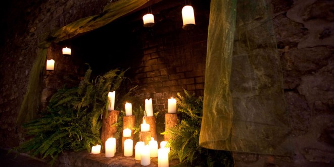 Lit candle display in a stone and brick fireplace, with green plants.