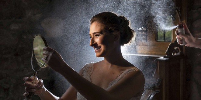 Bride looking into a handheld mirror, smiling, while light catches the hairspray being sprayed on her.