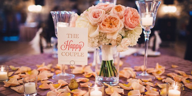 A small vase with a pink and white rose bouquet on a candle-lit table; "The Happy Couple" sign adjacent.