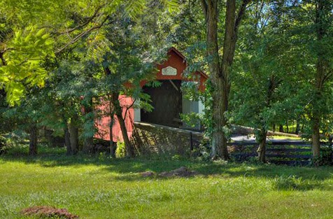 A red covered bridge tucked away behind green trees and a field of grass.