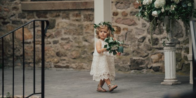 A flower girl holding a bouquet, walking near a stone building.