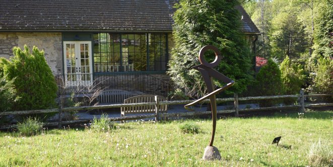 Justin Long's interpretive art statue in a field of grass, with an old building in the background.