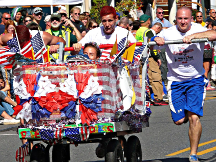 Two men running and pushing a red, white, and blue decorated wagon with someone in it.