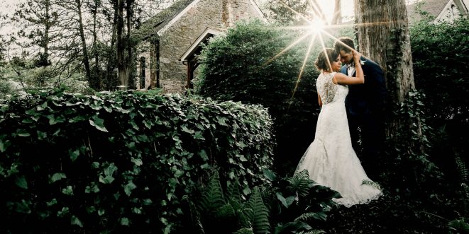 A bride and groom held in a loving embrace, surrounded by hedges and shrubs with sunlight peeking through.