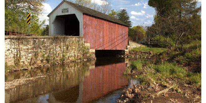 Red covered bridge situated on a lake, surrounded by greenery.