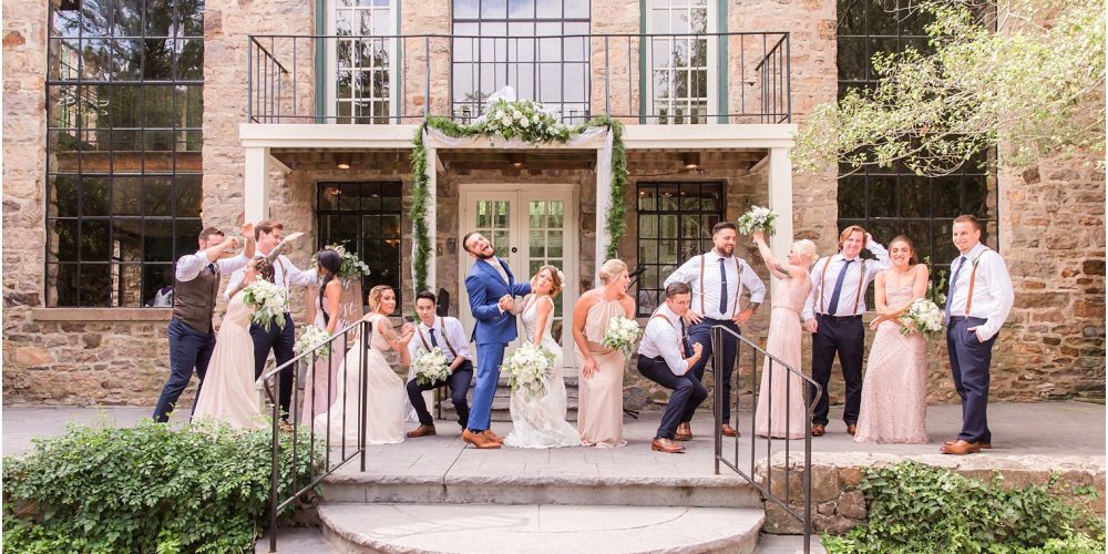 A bride, groom, and their bridal party in a line in front of the stone estate, making funny poses and faces.