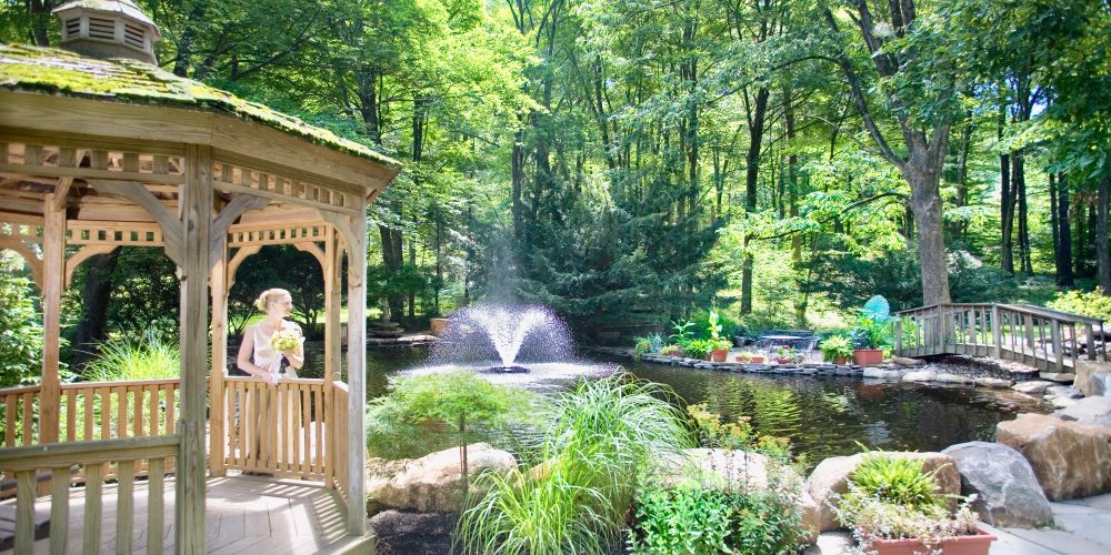 A bride standing under a gazebo with a view of a pond, fountain, and bridge in a wooded area.