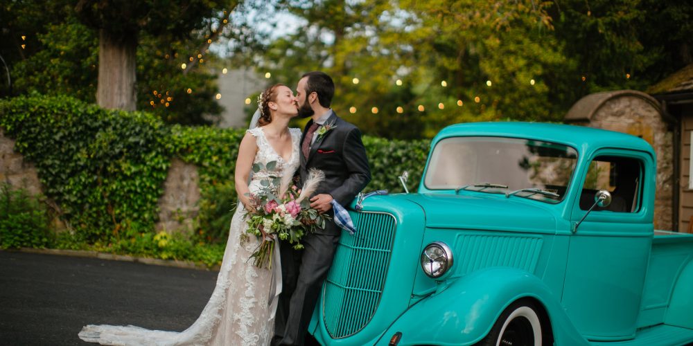 Bride and groom in front of an antique turquoise truck, kissing and holding a bouquet.