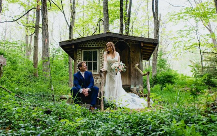 Bridge and groom pose in front of wooden structure in the woods, groom sitting, bridge standing and holding bouquet