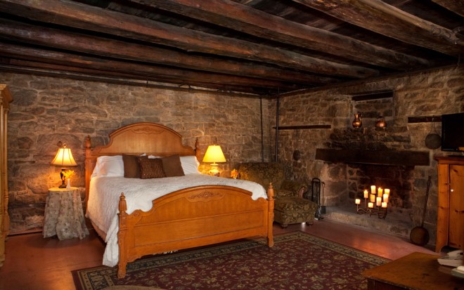 Photo of Room 11: The Honeymoon Suite is a large rustic room with stone walls and exposed wooden ceiling beams. A light wood bed is next to a large hearth.