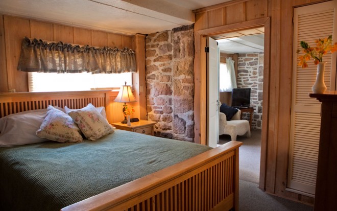 Photo of Room 15: A deluxe room with a bed with a light wood frame and green blanket. The walls are wood and stone and a door is open to an adjacent room.