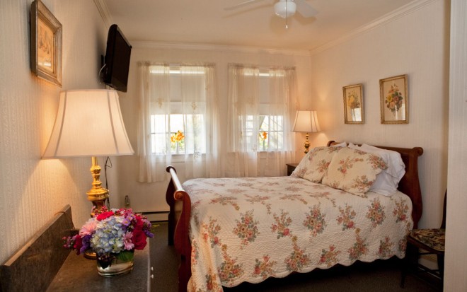 Photo of Room 5: A bright standard room with a dark wood bed and floral blanket, a lamp, and a TV on the wall